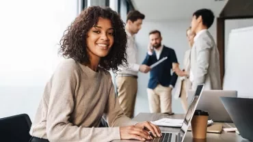 Persona sorridente con un laptop, sfondo con colleghi in ufficio.