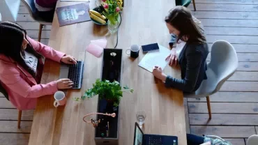 Due persone sedute a un tavolo in un ufficio, che lavorano su laptop e scrivono.