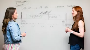Due persone discutono davanti a una lavagna con diagrammi tecnici.