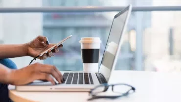 Mani che lavorano su un laptop e smartphone, tazza di caffè sul tavolo.