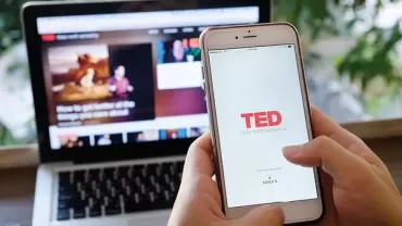 Persona che usa l'app TED su smartphone con laptop sullo sfondo.